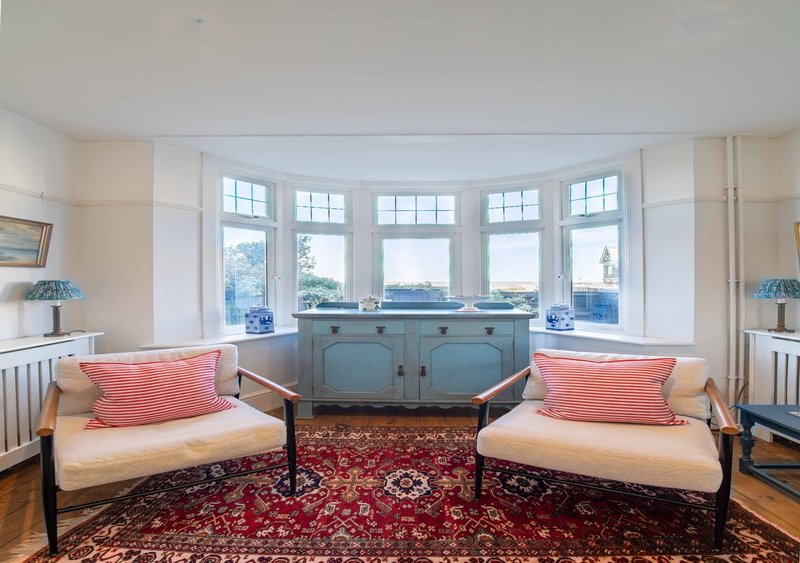 Bright living room with bay windows, two cream sofas with coral cushions, blue cabinet and red rug