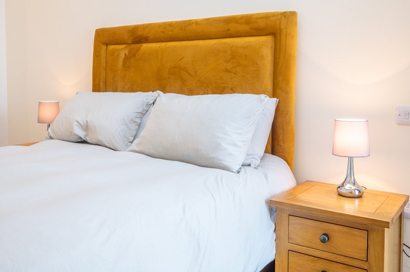 Bedroom with mustard-yellow upholstered headboard, white bedding, pillows, and wooden nightstand with lamp