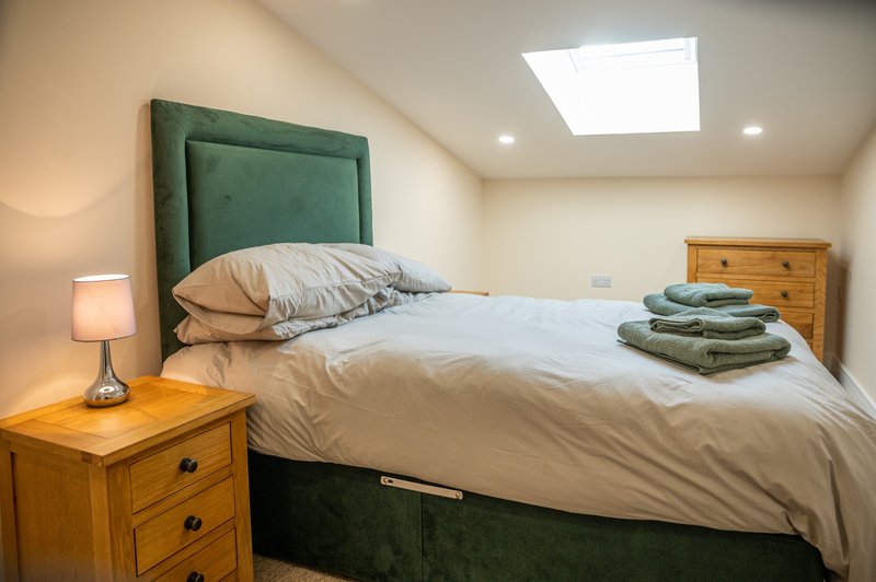 Bright bedroom with green headboard, wooden bed frame, bedside lamp, and folded towels on bed