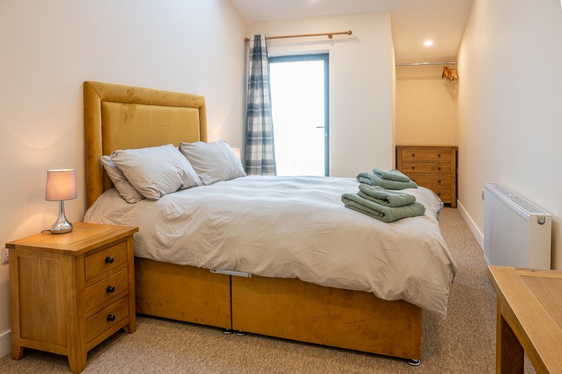 Bedroom with wooden double bed, mustard headboard, bedside table with lamp, and folded towels