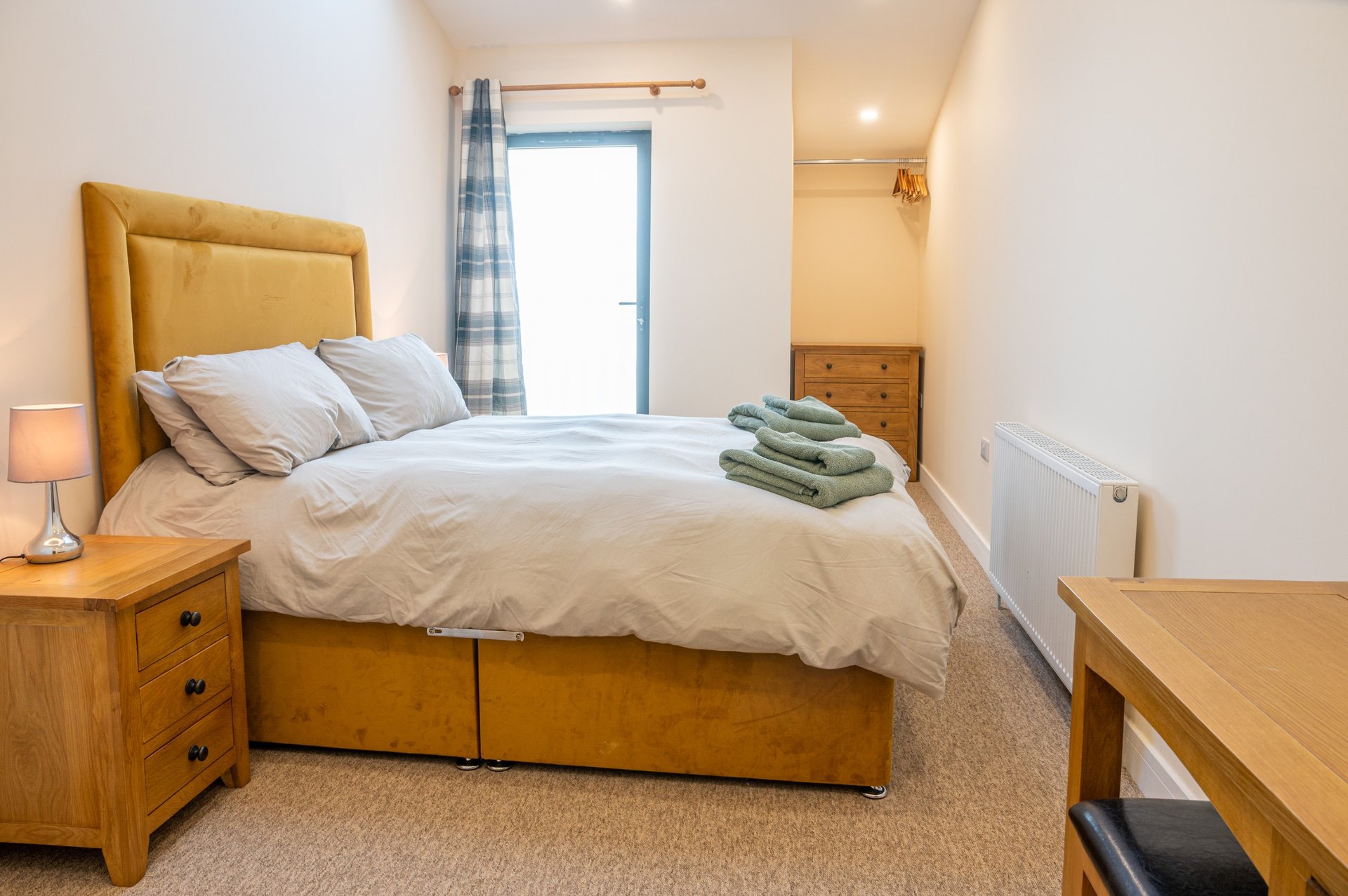 Bedroom with wooden bed frame, mustard headboard, folded towels, wooden furniture and door to patio