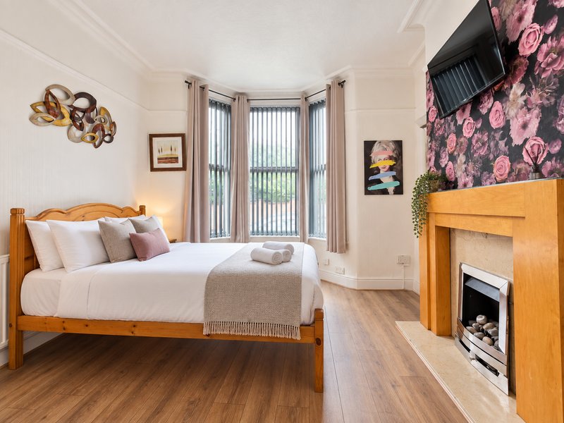Bright bedroom with wooden bed, fireplace, bay windows with black-out vertical blinds, and decorative wall art