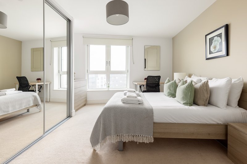 Modern bedroom with double bed, multiple pillows, mirrored wardrobe and bright windows