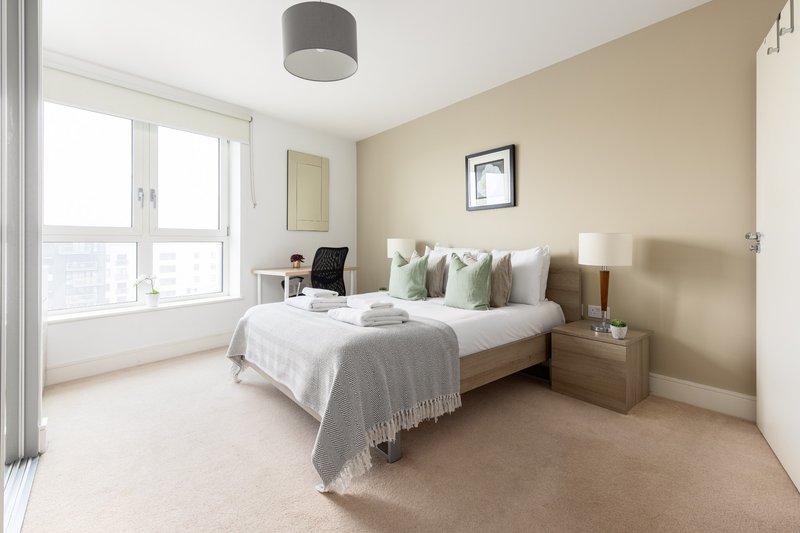 Bright modern bedroom with double bed, beige walls, large window and neutral furnishings