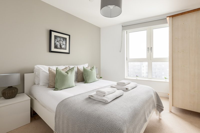 Bright bedroom with double bed, green cushions, white bedding, framed wall art and large window