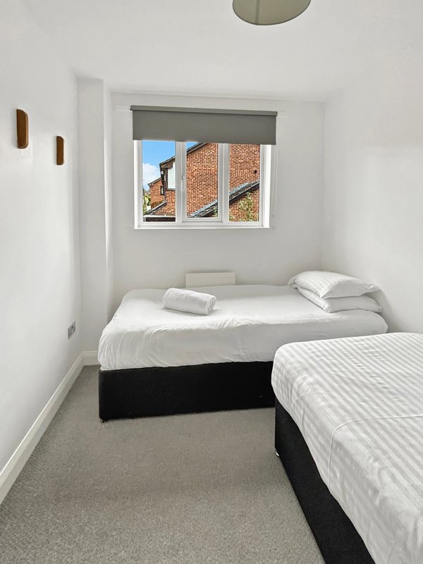 Small bedroom with two single beds, window with grey blind, white walls and grey carpet