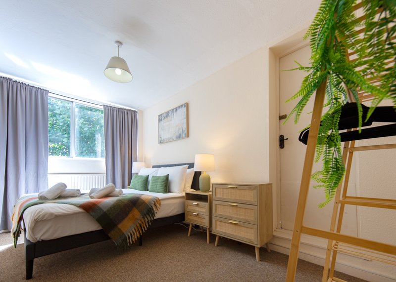 Bright bedroom with single bed, wooden nightstand, hanging plant, and large window with curtains