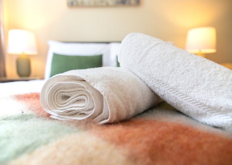 Bedroom with rolled towels on bed, green cushion, bedside lamps and warm lighting