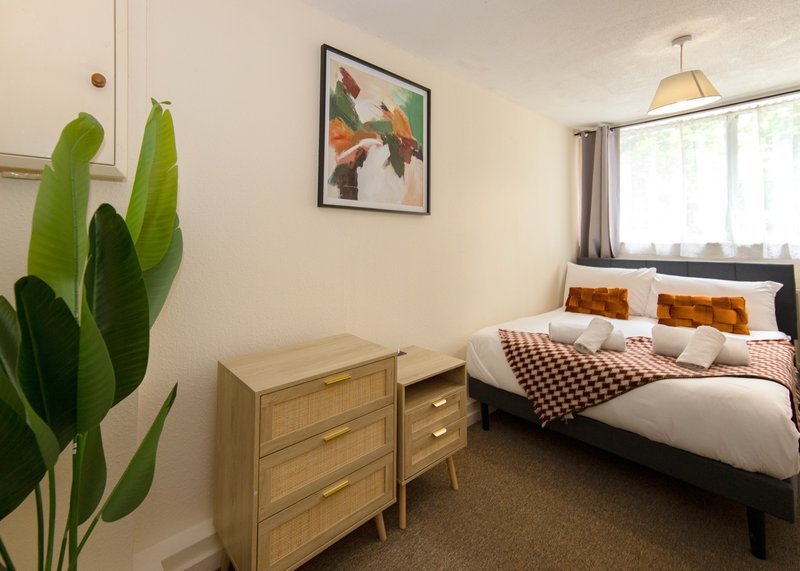 Bedroom with double bed, wooden dresser, green potted plant, framed wall art and window