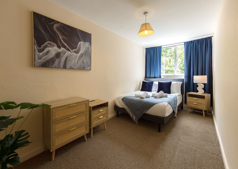 Bright bedroom with double bed, wooden dresser, wall art, blue curtains and bedside lamp