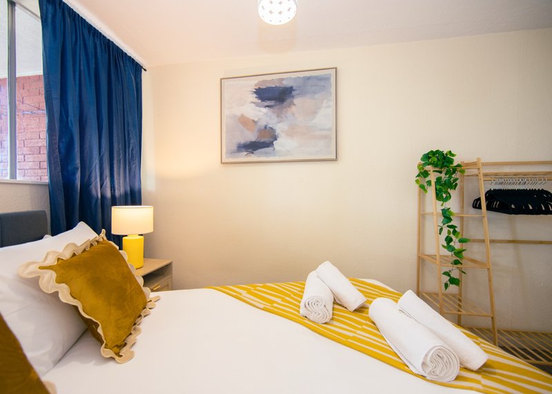 Bright bedroom with double bed, yellow bedding, mustard cushion, rolled towels, and blue curtains