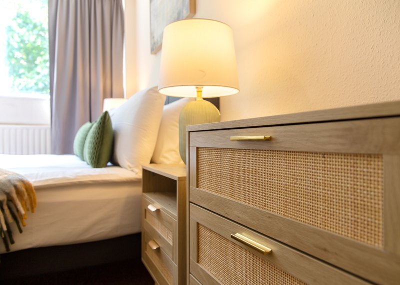 Bedroom with wooden bed frame, rattan-panel nightstand, table lamp, and window with grey curtains