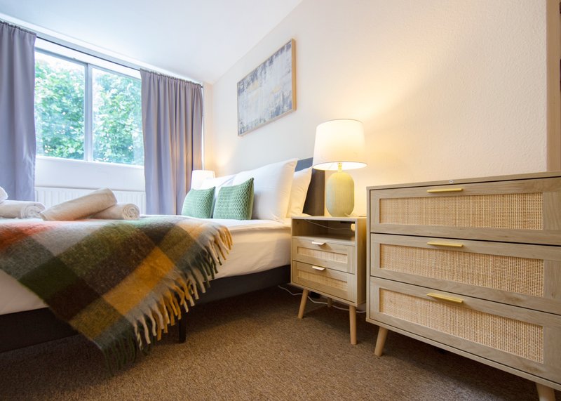 Bright bedroom with double bed, wooden dresser, bedside lamp, and large window with curtains