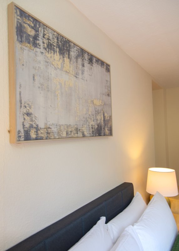 Bedroom corner with white pillows, black headboard, framed abstract wall art, and bedside lamp