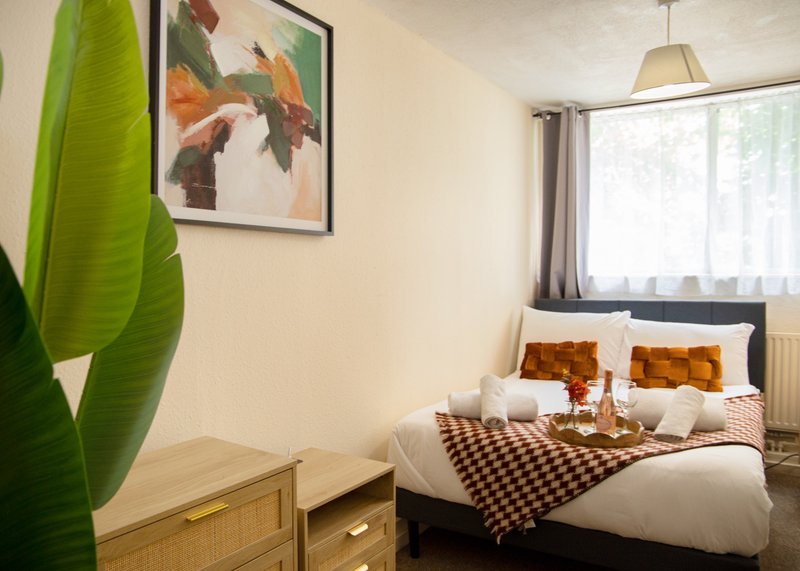 Bedroom with double bed, wooden furniture, large plant, framed artwork and window with curtains