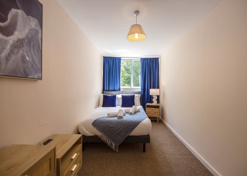 Compact bedroom with double bed, blue curtains, wooden desk, pendant light and window view