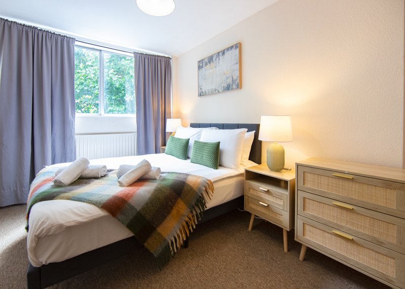 Bright bedroom with double bed, plaid throw, nightstands, dresser and window with curtains
