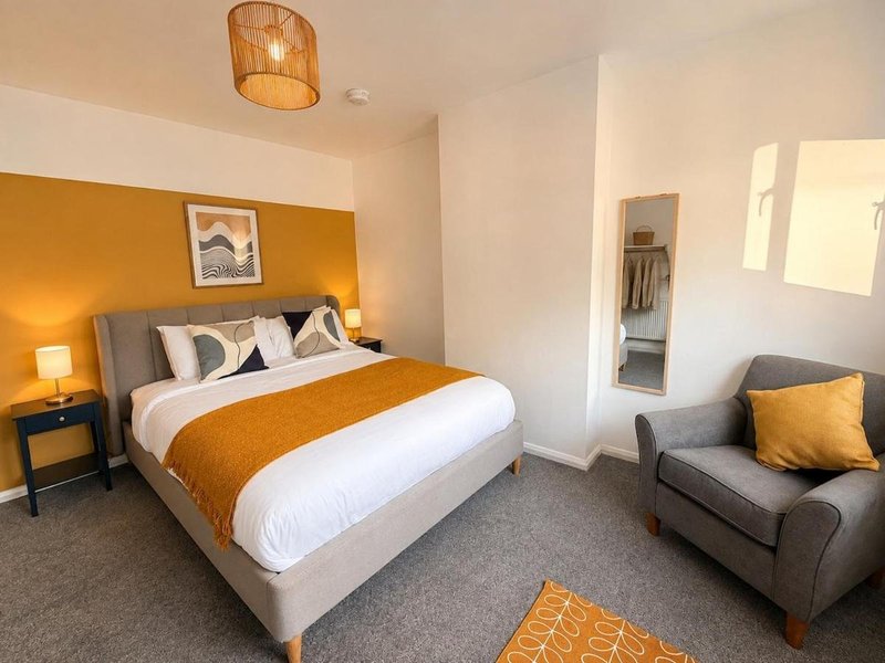 Modern bedroom with double bed, mustard headboard, armchair, and ensuite bathroom visible