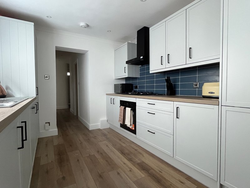 Modern galley kitchen with white cabinetry, black range hood, blue tile splashback and wooden flooring