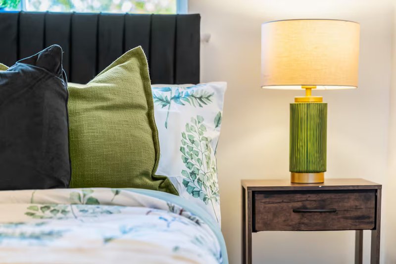 Bedroom detail with black headboard, green and white cushions, floral bedding, and wooden bedside table lamp