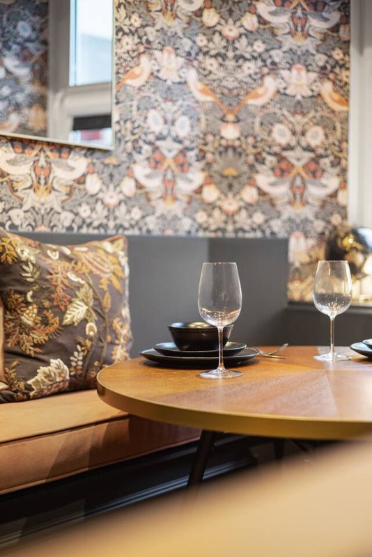 Dining table set with wine glasses, black plates, and gold decorative cushion against patterned wallpaper