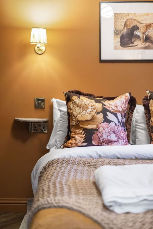 Warm ochre bedroom with double bed, decorative cushions, wall sconce lamp and framed artwork