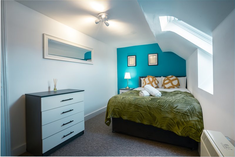 Bright bedroom with turquoise accent wall, double bed with patterned bedding, white dresser and skylight window