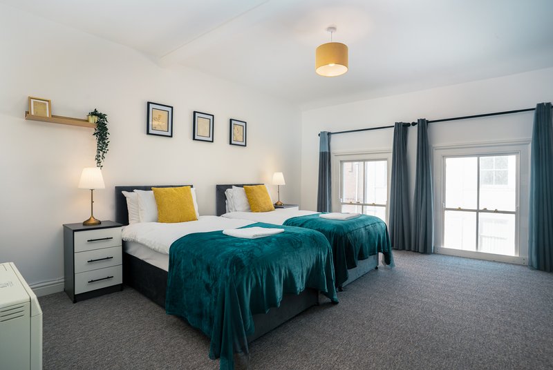 Modern bedroom with double beds, teal bedding, yellow and white pillows, wooden shelving and framed artwork