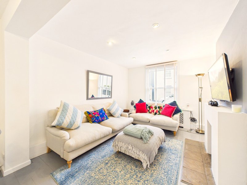 Bright living room with cream sofa, colorful cushions, footstool, windows and wall-mounted TV