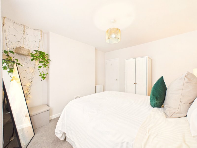 Bright modern bedroom with double bed, green and cream pillows, hanging plants, and wooden mirror