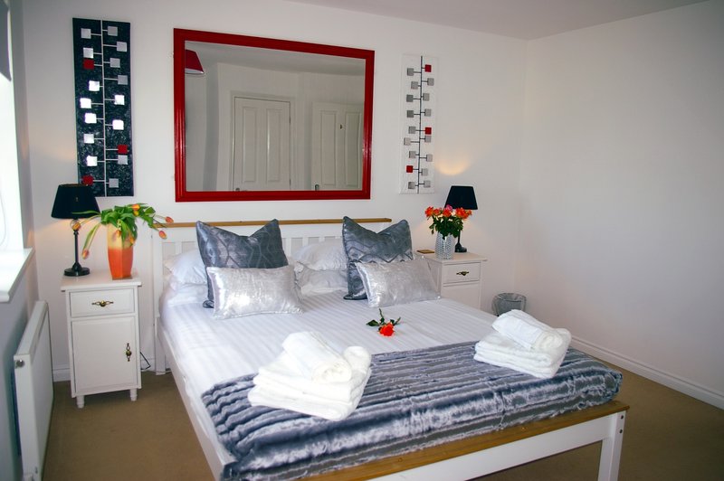 Bright bedroom with double bed, white bedding, red-framed mirror, bedside tables with lamps and flowers