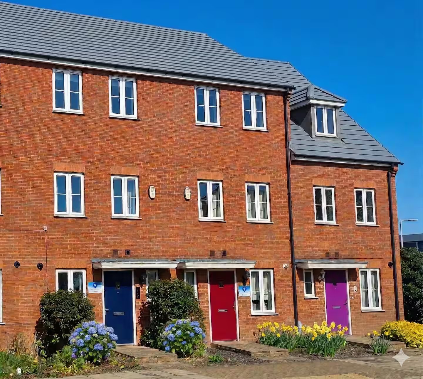 Modern brick townhouse with multiple colored doors, white windows, and front garden landscaping