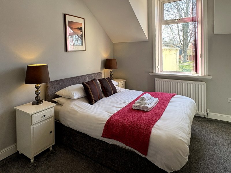 Bedroom with double bed, red throw, white bedside table, dark lamps and framed wall art