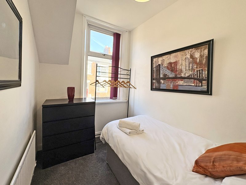 Compact bedroom with single bed, black dresser, window with burgundy curtains, framed cityscape print