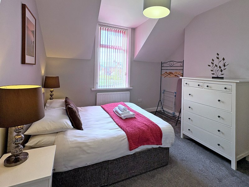 Bedroom with double bed, white dresser, lamp, and window with pink walls