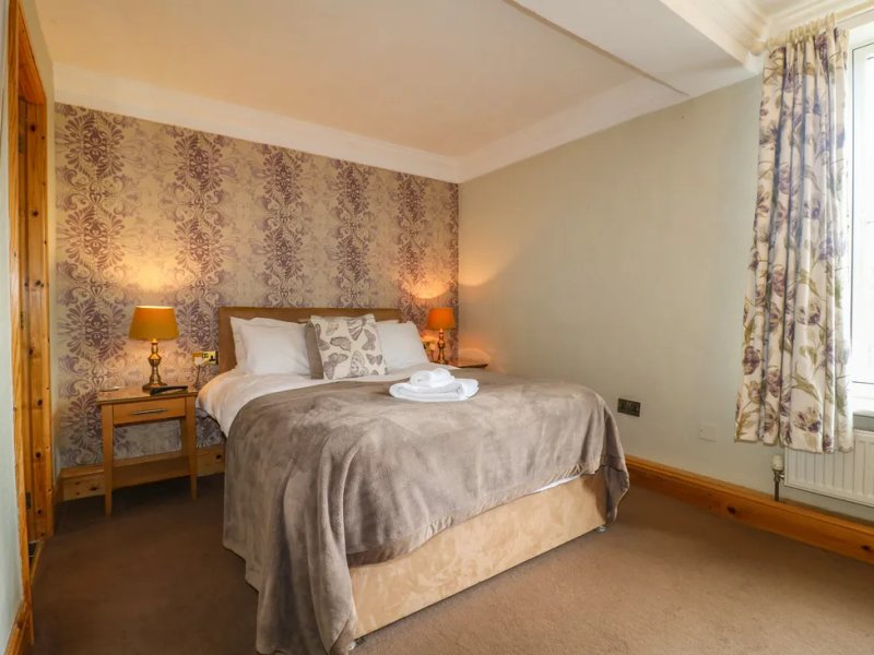 Bedroom with double bed, patterned wallpaper, wooden bedside tables, and floral curtains