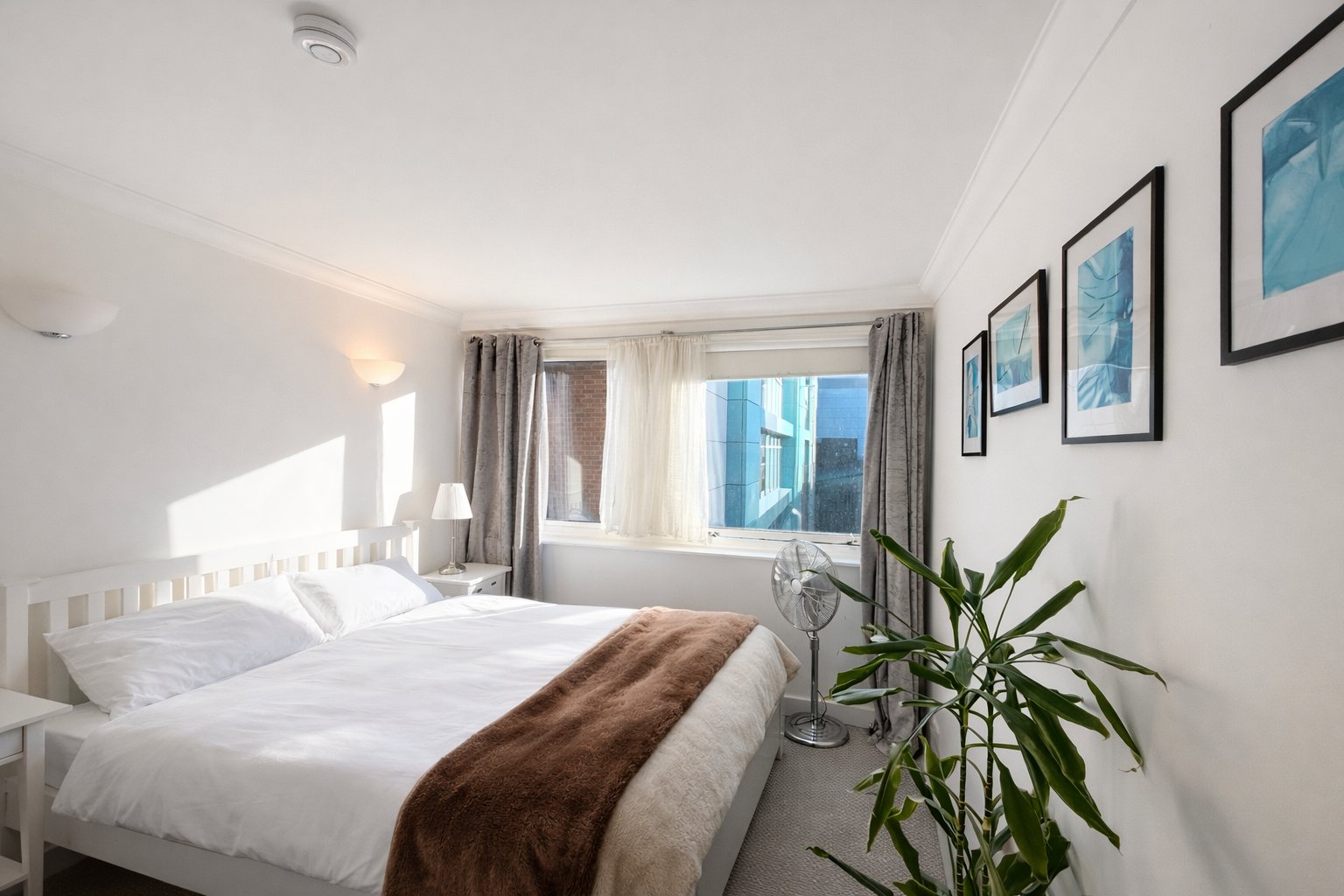 Bright modern bedroom with double bed, white bedding, brown throw, plant and framed wall art