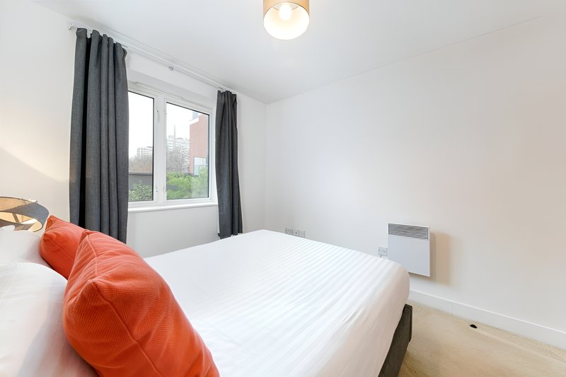 Bright bedroom with double bed, orange cushions, window with grey curtains, wooden floor