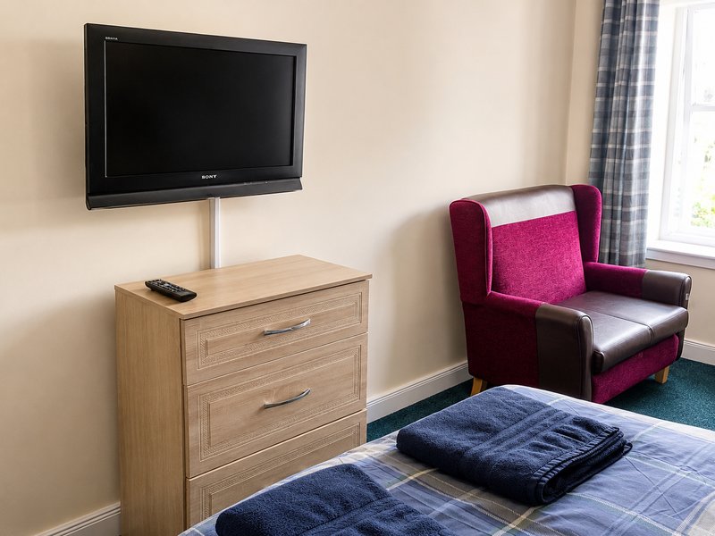Bedroom with wall-mounted TV, wooden chest of drawers, burgundy armchair and blue bedding