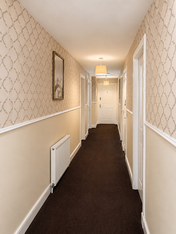 Long hallway with patterned wallpaper, white trim, dark carpet and ceiling light