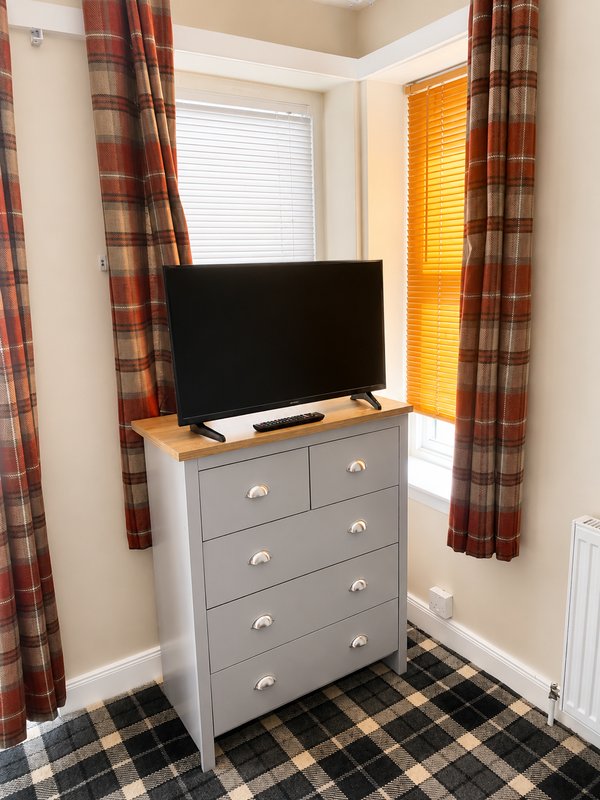 Bedroom corner with grey chest of drawers, flat-screen TV, plaid curtains and checked floor