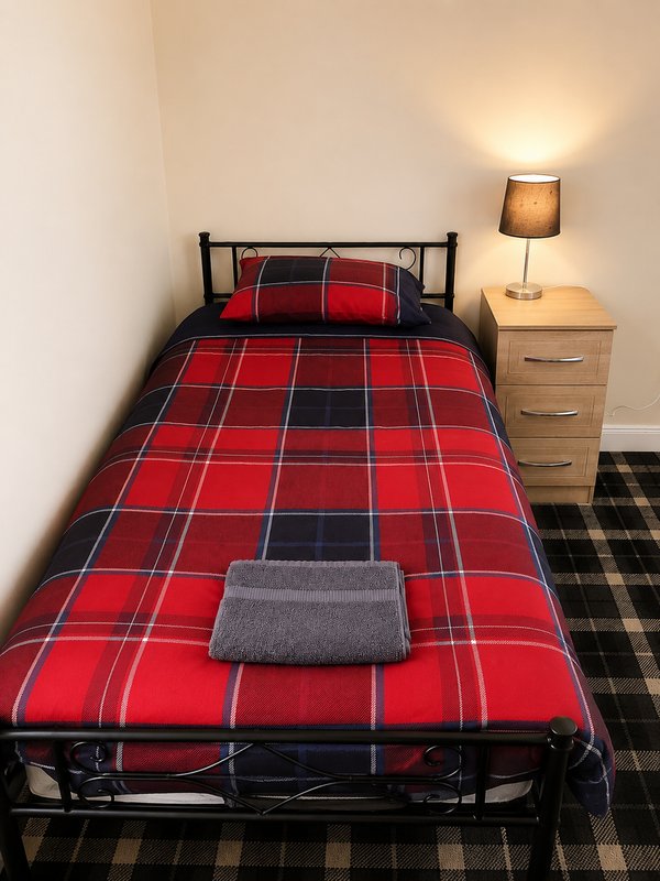 Single bedroom with red tartan bedding, metal frame bed, wooden nightstand and lamp