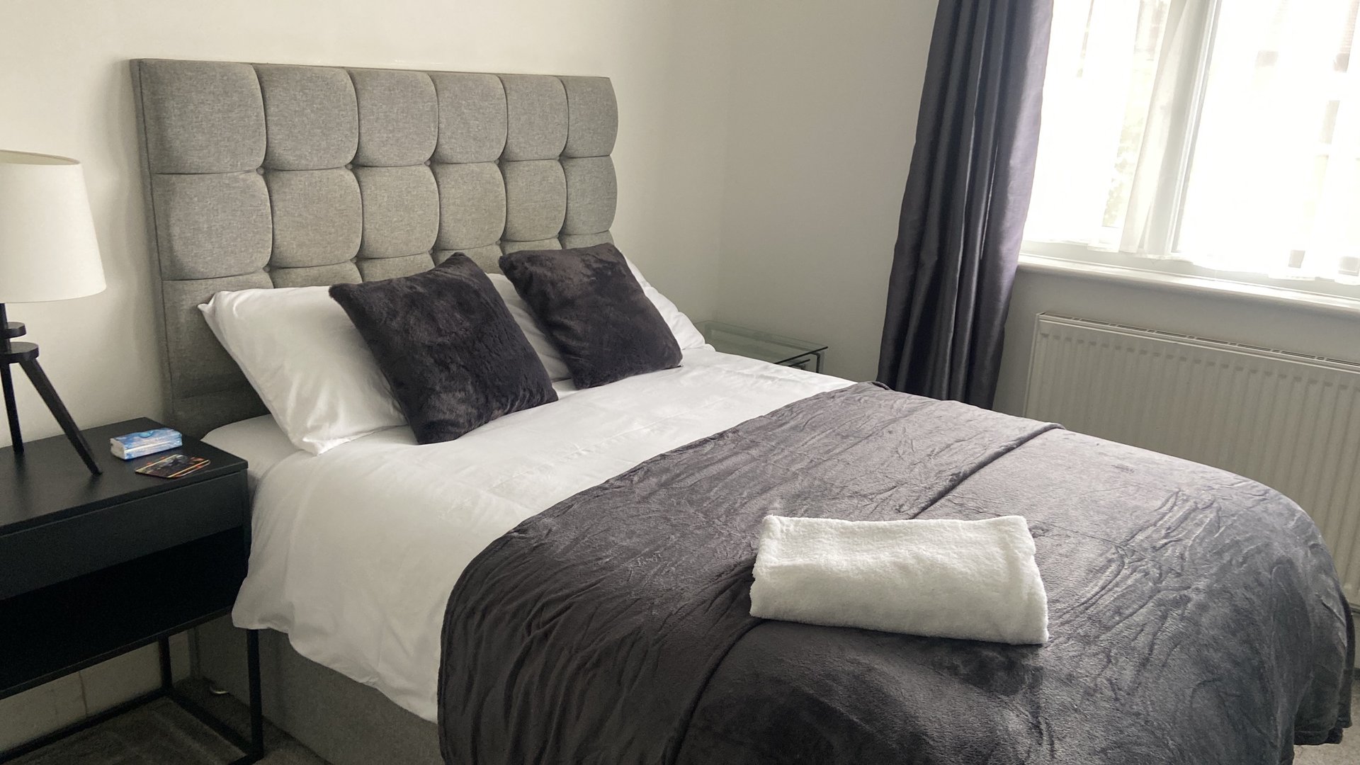 Modern bedroom with grey upholstered headboard, white and grey bedding, and window with blue curtains