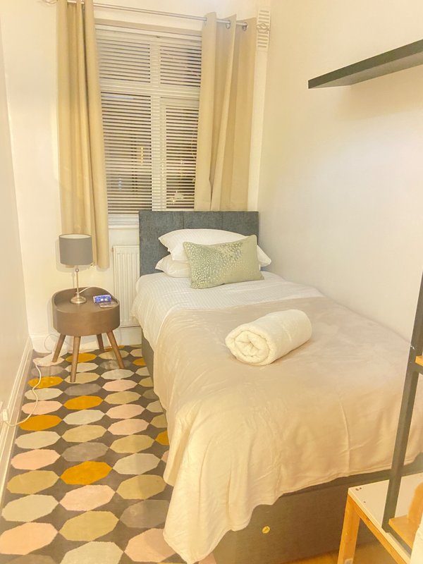 Compact bedroom with single bed, grey headboard, cream bedding, round wooden side table and patterned floor