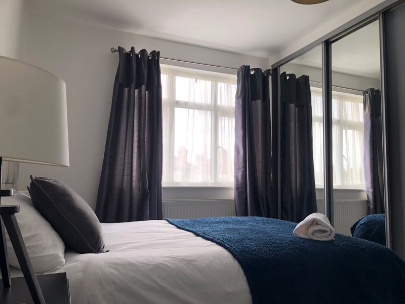Bright bedroom with double bed, blue throw blanket, dark curtains and mirrored wardrobe