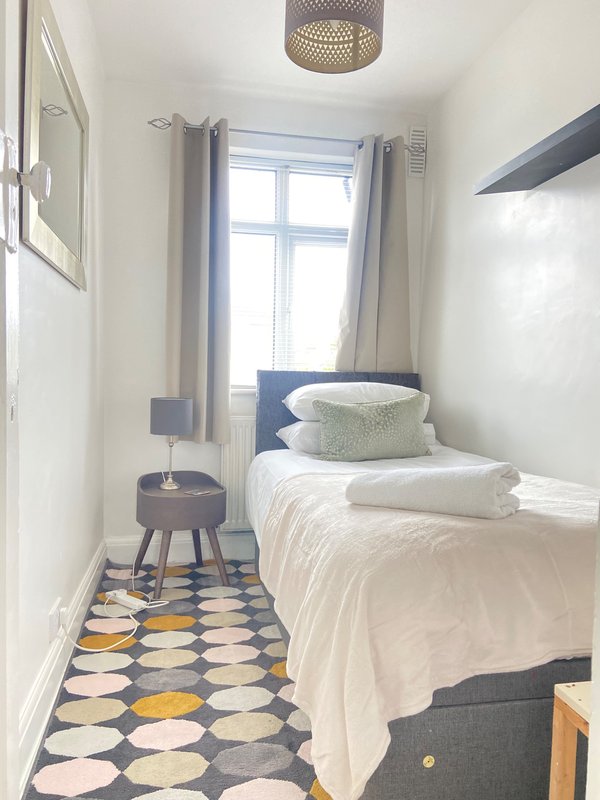 Small bedroom with single bed, white bedding, pillows, window with grey curtains, geometric floor rug
