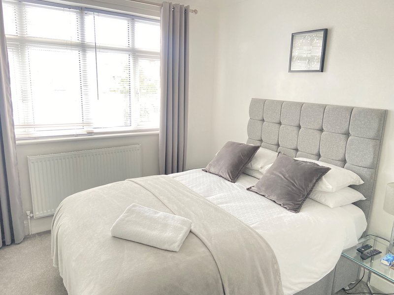 Bright bedroom with double bed, grey upholstered headboard, white bedding and large window with curtains
