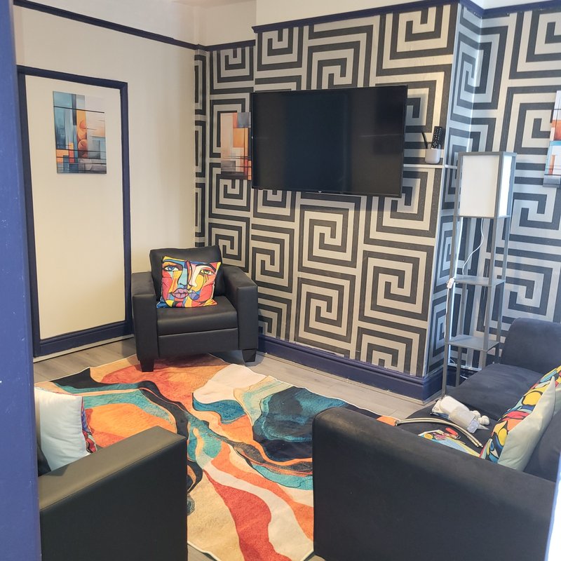 Living room with geometric wallpaper, TV, black armchair, colorful area rug and seating.