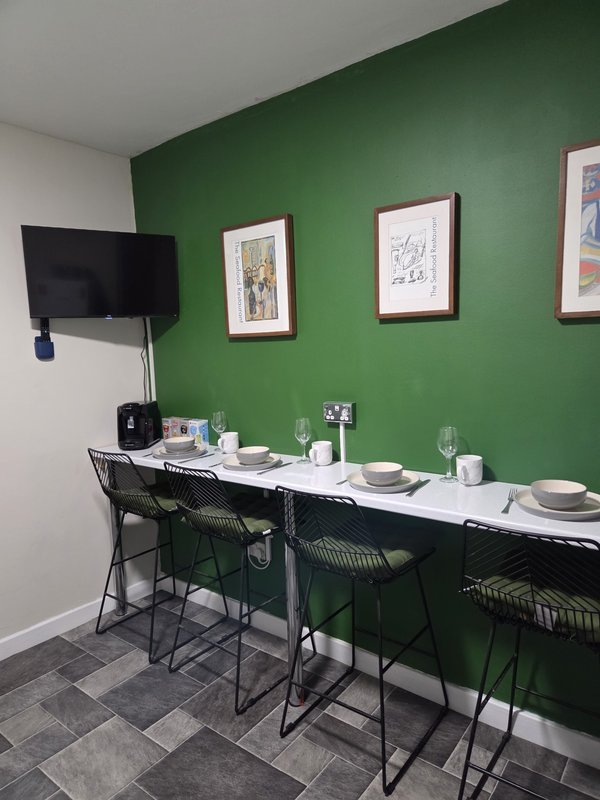 Dining area with long table, black chairs, green wall, framed artwork, and mounted television.
