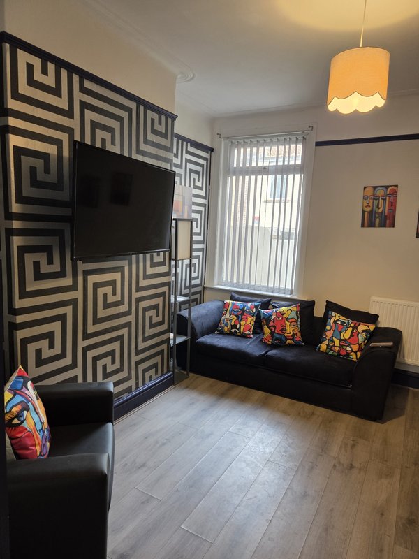 Living room with black sofa, geometric wall art, flat-screen TV, pendant lamp, and colorful cushions